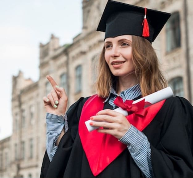 A female student in a graduation robe is posing in front of the college building.