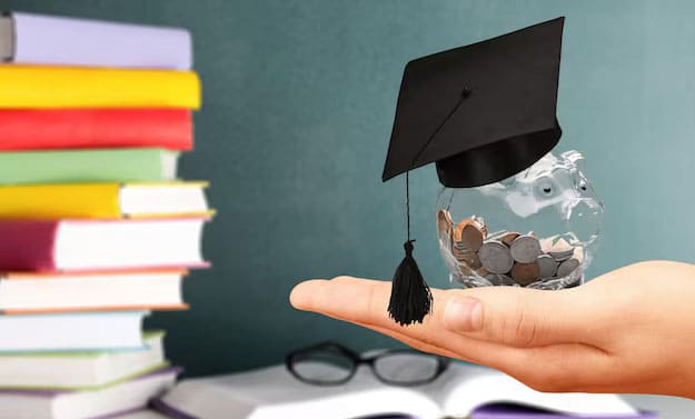 Alongside books, someone is holding a piggy bank partially covered with a graduation hat in their left palm.